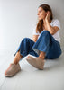 A woman sits on a white wooden floor against a white wall, wearing beige slip-on slippers with red stitching, blue jeans, and a white t-shirt.