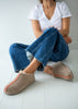 A person sitting on a white floor wearing taupe suede slip-on slippers with a pink stitched accent line, blue jeans, and a white t-shirt.