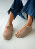 A person wearing beige slip-on sheepskin slipper boots with a pink patterned central stripe, paired with frayed blue jeans, sitting on a white wooden floor.