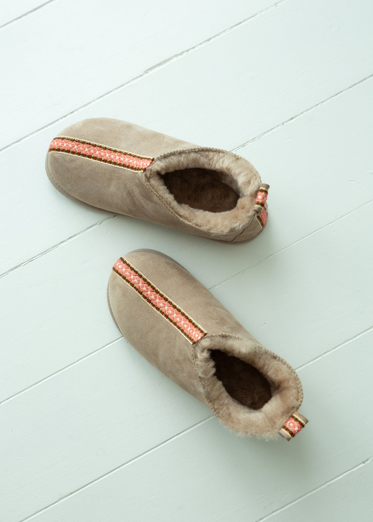 A pair of taupe suede slipper boots with a soft beige sheepskin lining and a decorative coral-orange patterned stripe, displayed on a light blue wooden plank floor.