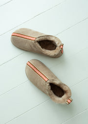 A pair of taupe suede slipper boots with a soft beige sheepskin lining and a decorative coral-orange patterned stripe, displayed on a light blue wooden plank floor.