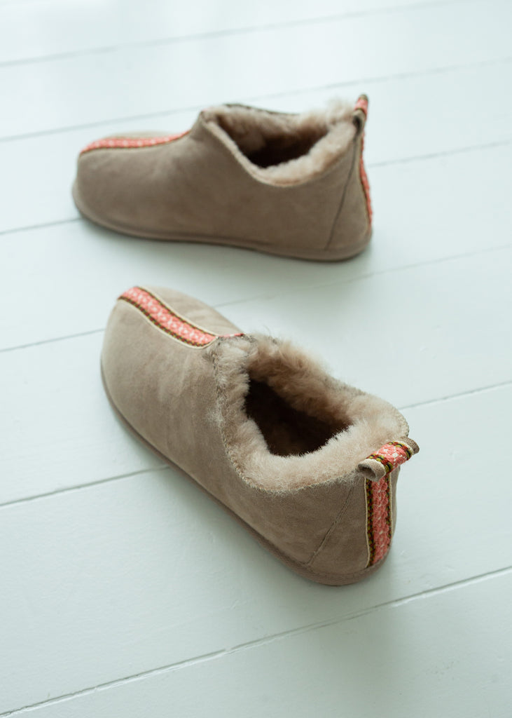 A pair of slip-on slippers with a taupe suede-like exterior, plush cream shearling lining, and a thin orange-red decorative stripe along the side, displayed on a white wooden plank floor.