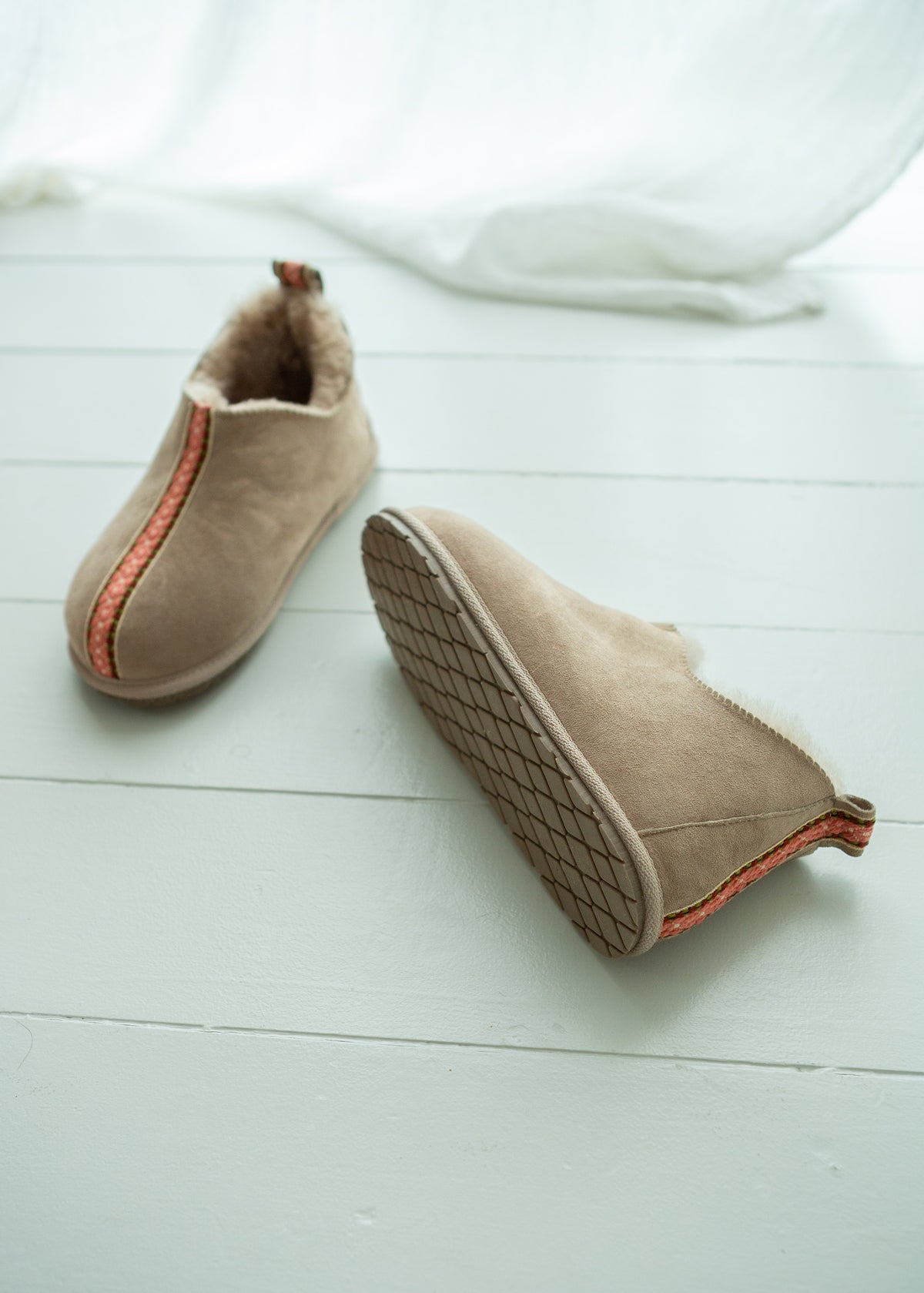 A pair of beige sheepskin booties with a soft cream lining and a thin orange-red stripe detail, displayed on a white wooden floor with a white fabric drape in the background.