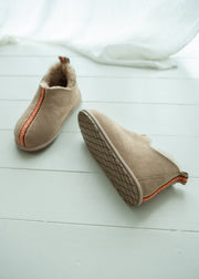 A pair of beige sheepskin booties with a soft cream lining and a thin orange-red stripe detail, displayed on a white wooden floor with a white fabric drape in the background.