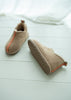 A pair of beige sheepskin booties with a soft cream lining and a thin orange-red stripe detail, displayed on a white wooden floor with a white fabric drape in the background.