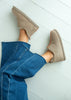 A top-down view of a person wearing taupe suede slip-on slippers with frayed edges, paired with blue jeans, on a light gray wood plank floor.