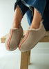 A person wearing beige slip-on sheepskin slippers with a pink patterned stripe down the front, sitting on a wooden bench. The slippers have a soft fuzzy lining and are shown with frayed-hem blue jeans against a bright, light background.