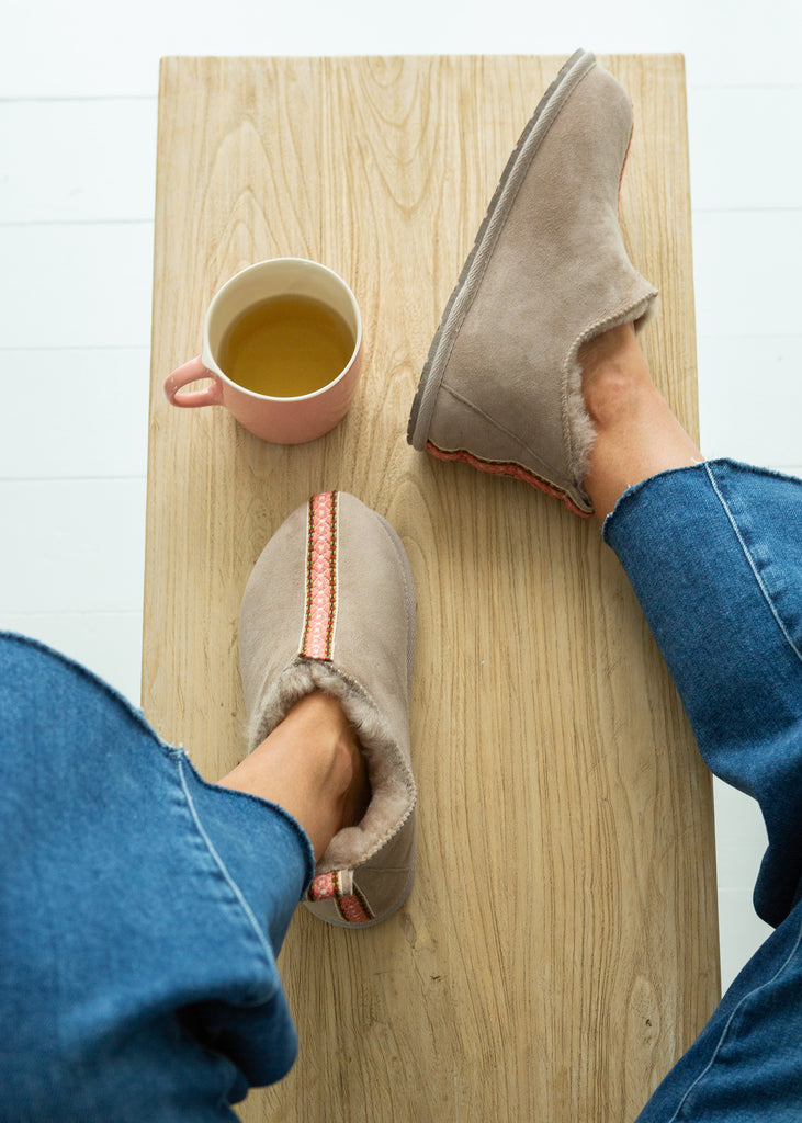 A top-down view of a person wearing cozy taupe slip-on sheepskin slipper boots with a red stripe detail, sitting on a light wood bench with a pink mug of tea nearby.