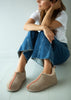 A woman sits on a light gray floor wearing taupe slip-on slippers with a coral-pink stitched detail and blue jeans.