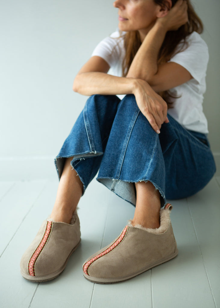 A woman sits on a light gray floor wearing taupe slip-on slippers with a coral-pink stitched detail and blue jeans.