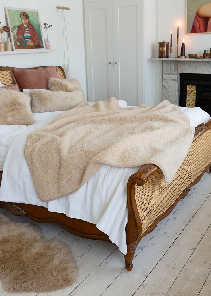 A cozy bedroom scene featuring a bed styled with a plush, soft beige sheepskin or faux-shearling throw blanket draped over white bedding, set in a warm, rustic bedroom with a cane and wood frame.