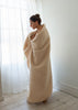 A woman wrapped in a plush, cream-colored teddy blanket standing in a bright room with white walls, light wood floors, and sheer white curtains.
