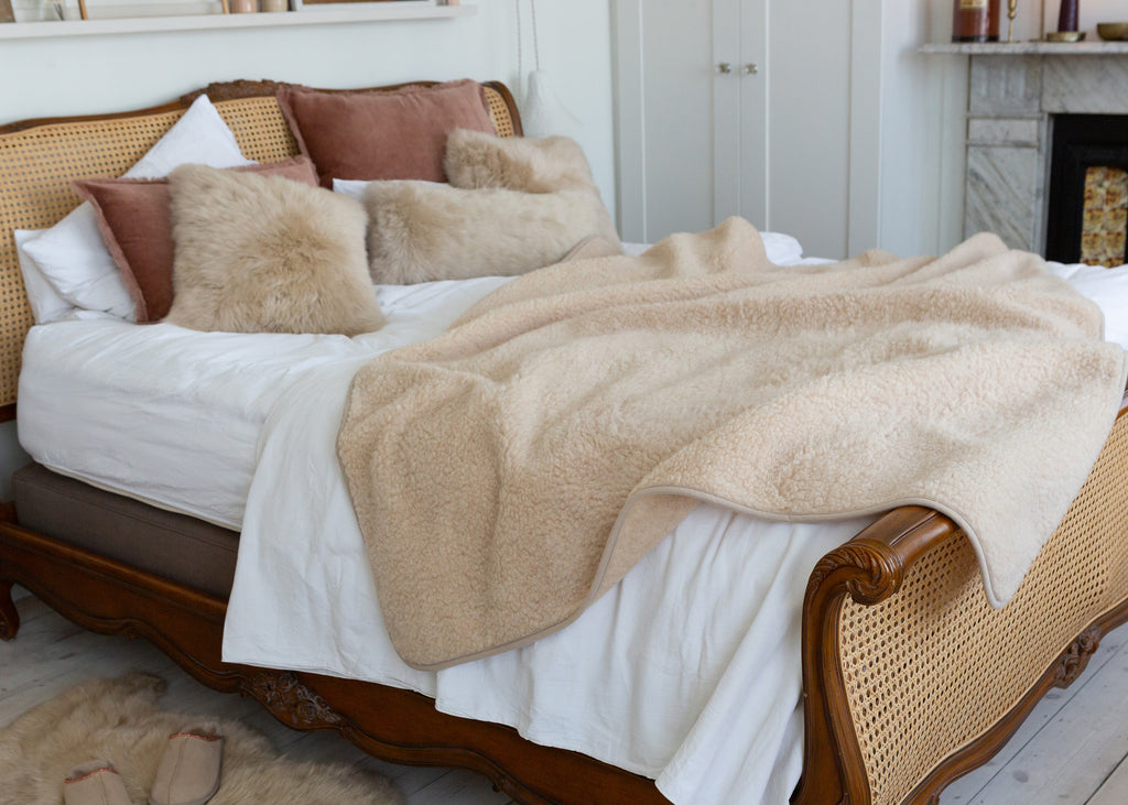 A cozy bedroom scene featuring a plush beige sherpa throw blanket draped over a bed with white bedding, styled with brown and beige pillows on a wooden cane bed frame.