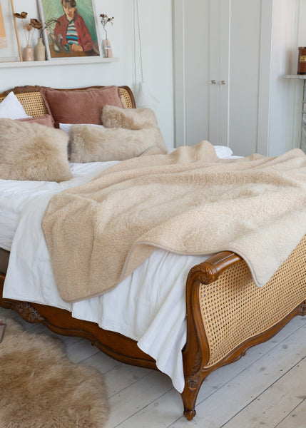 A cozy bedroom scene featuring a bed styled with a soft beige sherpa throw blanket, white bedding, and textured beige pillows on a wooden bed frame with woven cane panels.