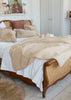 A cozy bedroom scene featuring a bed styled with a soft beige sherpa throw blanket, white bedding, and textured beige pillows on a wooden bed frame with woven cane panels.