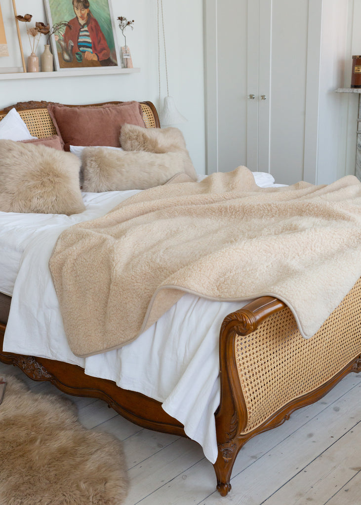 A cozy bedroom scene featuring a bed styled with a soft beige sherpa throw blanket, white bedding, and textured beige pillows on a wooden bed frame with woven cane panels.