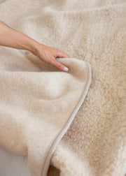 A close-up of a person's hand lifting a corner of a soft, cream-colored sheepskin or faux-sheepskin blanket to show its thick, plush texture.