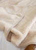 A close-up of a soft, plush fleece blanket in a warm cream or light beige color, draped over a white surface with a wooden furniture leg visible in the corner.
