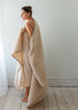 A woman wrapped in a plush cream-colored fleece blanket or throw, standing barefoot in a minimalist room with light wood floors and a white wall.
