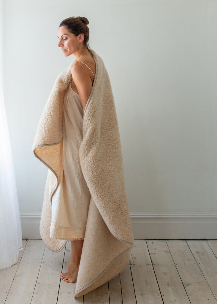 A woman wrapped in a plush cream-colored fleece blanket or throw, standing barefoot in a minimalist room with light wood floors and a white wall.
