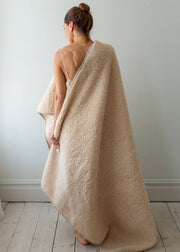A woman stands with her back to the camera, wrapped in a large, plush, cream-colored teddy blanket or throw in a minimalist room with light gray walls and white wooden floors.