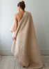 A woman stands with her back to the camera, wrapped in a large, plush, cream-colored teddy blanket or throw in a minimalist room with light gray walls and white wooden floors.