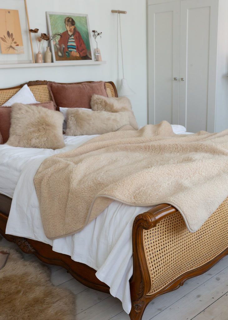 A cozy bedroom scene featuring a bed styled with a plush beige faux fur throw blanket, matching beige pillows, white bedding, and a wooden cane and wood frame bed.