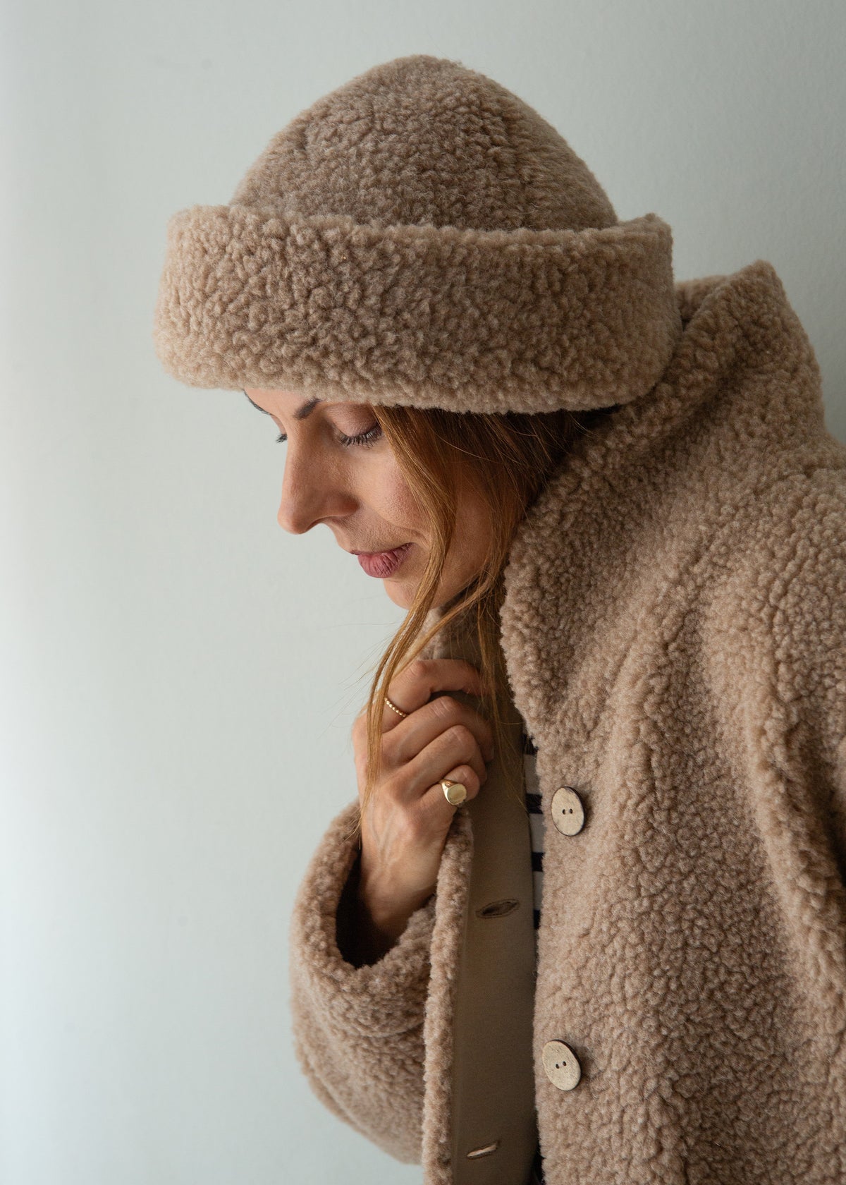 A woman wearing a cozy taupe-brown merino fleece beanie and matching fleece jacket, shown in a close-up profile against a plain light gray background.