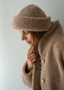 A woman wearing a cozy taupe-brown merino fleece beanie and matching fleece jacket, shown in a close-up profile against a plain light gray background.