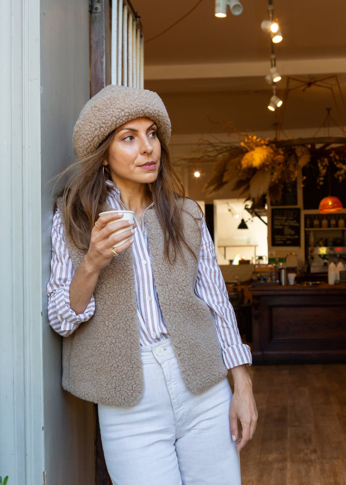 A woman wearing a beige merino beanie, a taupe merino gilet over a white and navy striped button-down shirt, and white jeans, leaning against a wall inside a cozy cafe.
