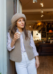 A woman wearing a beige merino beanie, a taupe merino gilet over a white and navy striped button-down shirt, and white jeans, leaning against a wall inside a cozy cafe.