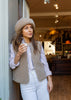 A woman wearing a beige merino beanie, a taupe merino gilet over a white and navy striped button-down shirt, and white jeans, leaning against a wall inside a cozy cafe.