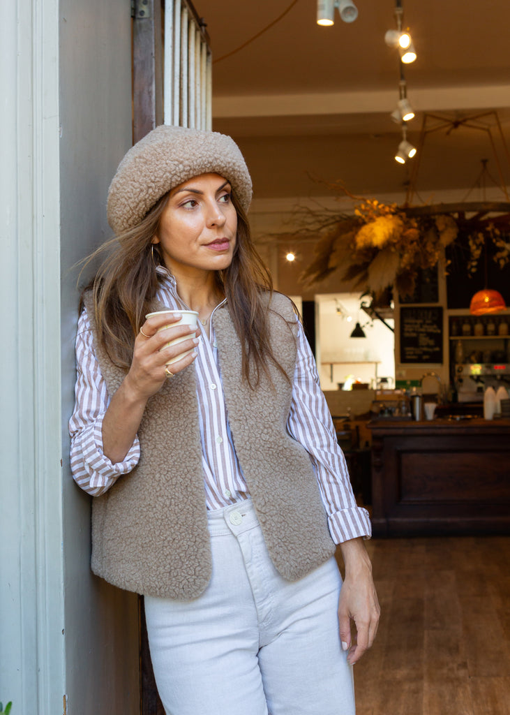A woman wearing a beige merino beanie, a taupe merino gilet over a white and navy striped button-down shirt, and white jeans, leaning against a wall inside a cozy cafe.