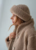 A woman in profile wearing a cozy taupe merino fleece beanie and matching fleece jacket against a plain light gray background.