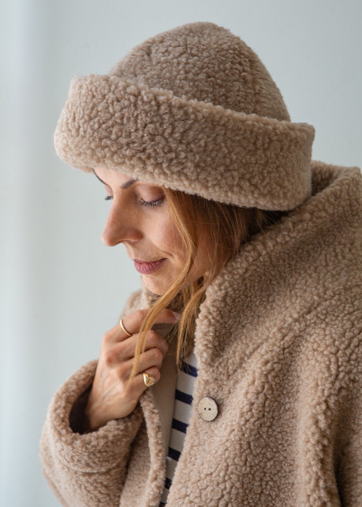 A woman wearing a cozy taupe-brown merino fleece beanie hat and a matching fleece jacket, looking down in a soft, indoor setting.