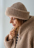 A woman wearing a cozy taupe-brown merino fleece beanie hat and a matching fleece jacket, looking down in a soft, indoor setting.