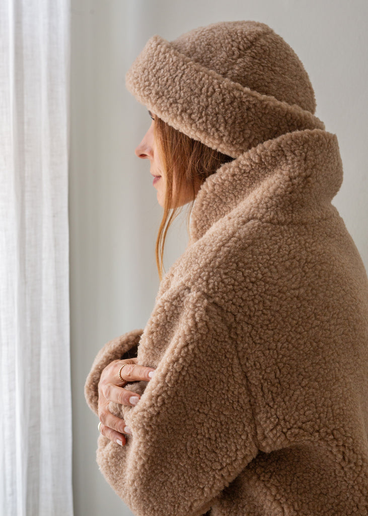 A person shown in side profile wearing a cozy camel brown merino coat with a matching merino hat, standing near a white curtain.