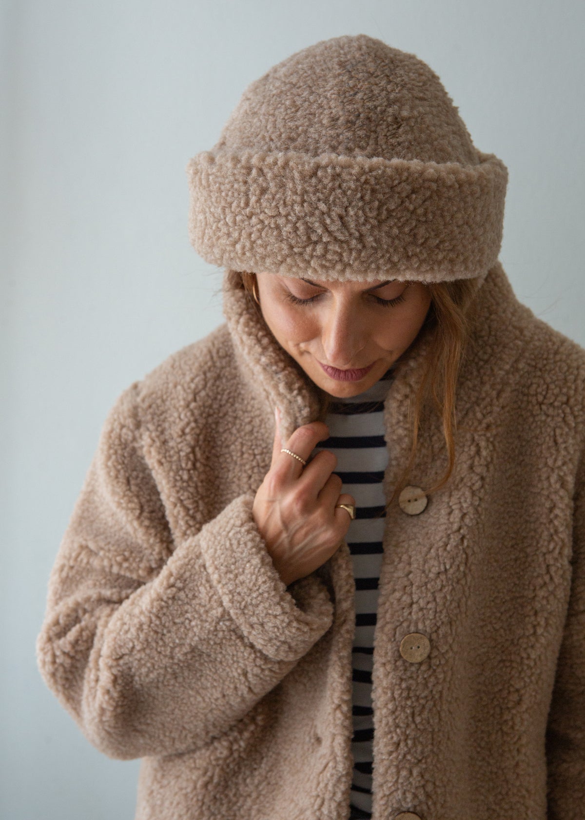 A woman wearing a cozy beige sherpa merino beanie and matching fleece jacket over a black and white striped shirt, looking down against a light gray background.