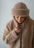A woman wearing a cozy beige sherpa merino beanie and matching fleece jacket over a black and white striped shirt, looking down against a light gray background.