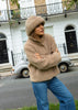 A woman wearing a beige teddy fleece merino jacket and matching beanie hat, posing outdoors in front of a white building with a vintage car in the background.
