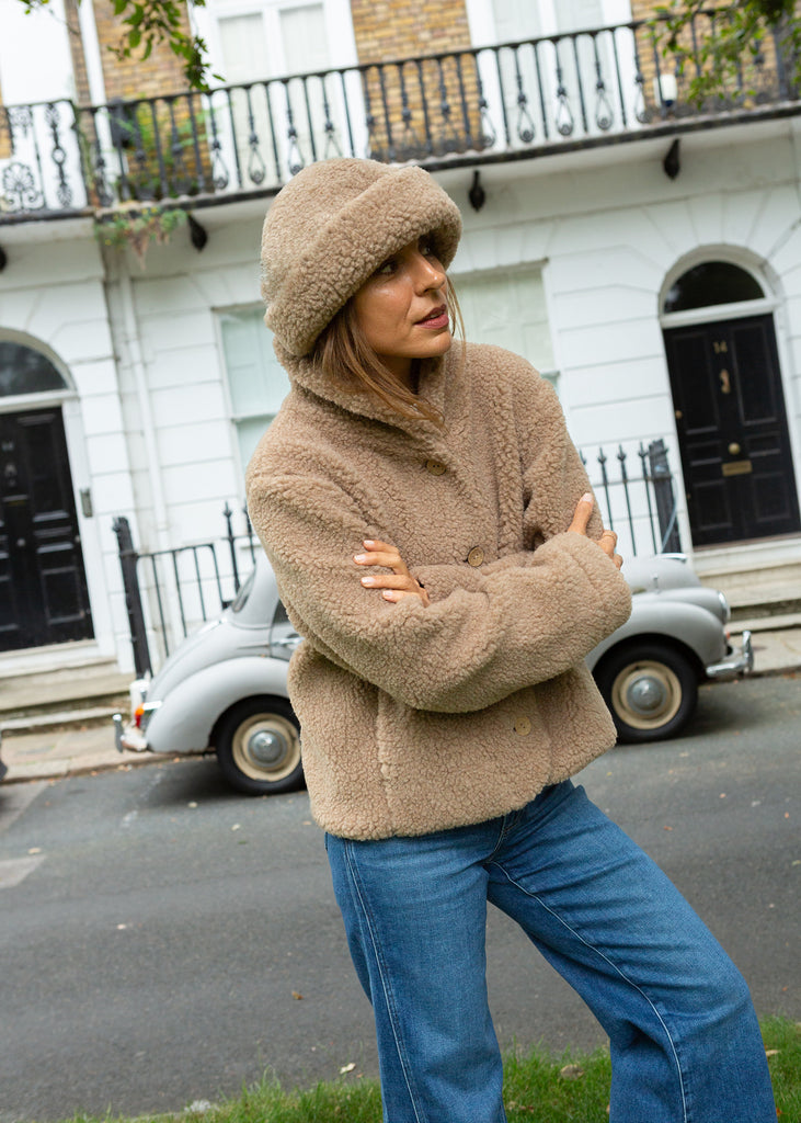 A woman wearing a beige teddy fleece merino jacket and matching beanie hat, posing outdoors in front of a white building with a vintage car in the background.