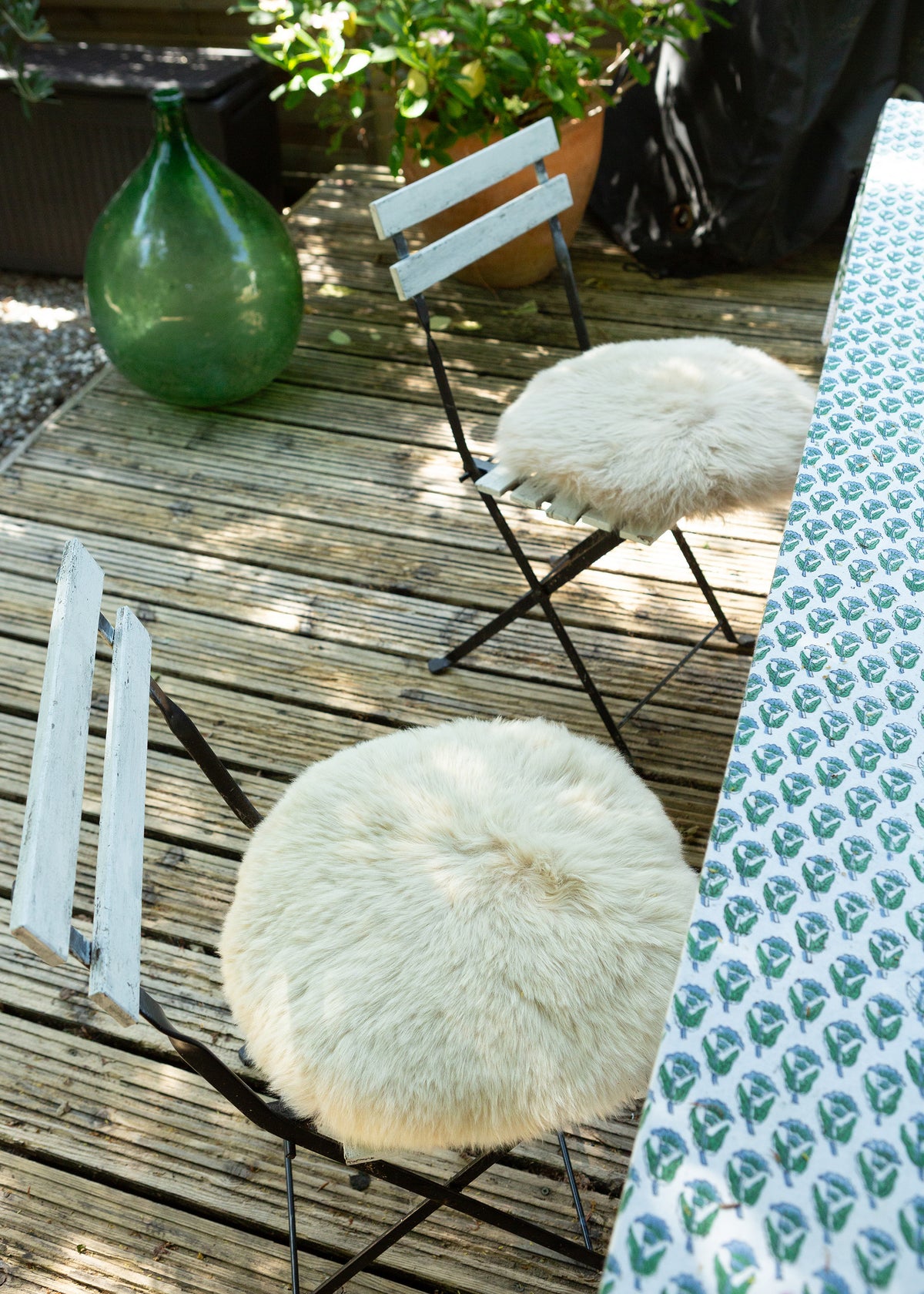 Two outdoor folding chairs with round, fluffy cream sheepskin seats on a wooden deck, next to a patterned tablecloth.