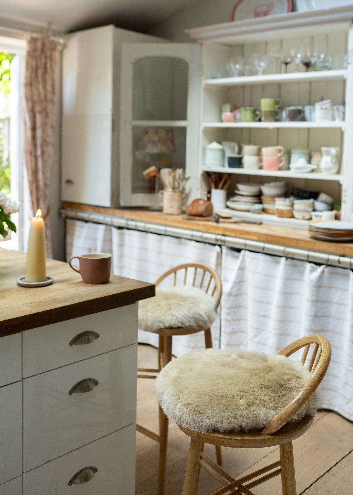 A cozy kitchen interior with wooden chairs featuring fluffy cream sheepskin seat pads, a white kitchen island, and open shelving filled with dishes and glassware.
