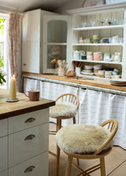 A cozy kitchen interior with wooden chairs featuring fluffy cream sheepskin seat pads, a white kitchen island, and open shelving filled with dishes and glassware.