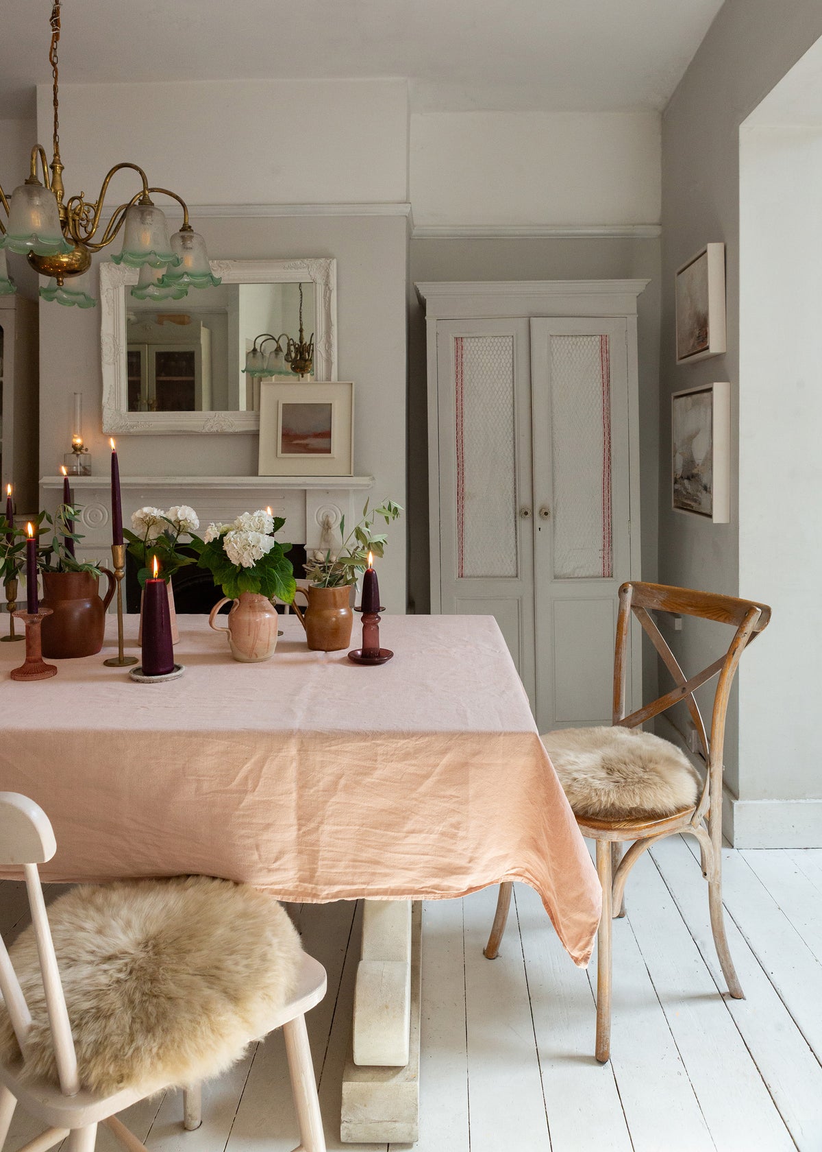 A dining room scene with a pale blush pink tablecloth on a wooden table, surrounded by wooden chairs with beige sheepskin seat pads. The room features light gray walls, a white fireplace with a mirror, and a brass chandelier.