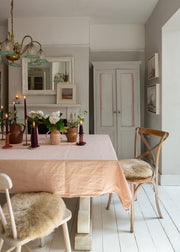 A dining room scene with a pale blush pink tablecloth on a wooden table, surrounded by wooden chairs with beige sheepskin seat pads. The room features light gray walls, a white fireplace with a mirror, and a brass chandelier.