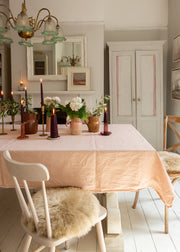 A dining room scene with a pale blush pink tablecloth on a wooden table, surrounded by wooden chairs with beige sheepskin seat pads. The room features light gray walls, a white fireplace with a mirror, and a brass chandelier.
