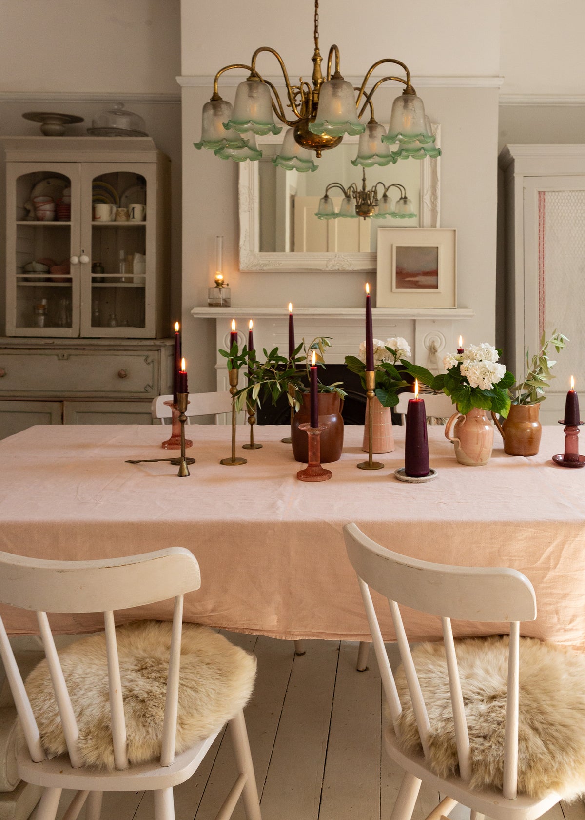A dining room scene with a pale blush pink tablecloth on a wooden table, surrounded by wooden chairs with beige sheepskin seat pads. The room features light gray walls, a white fireplace with a mirror, and a brass chandelier.
