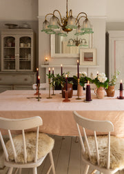 A dining room scene with a pale blush pink tablecloth on a wooden table, surrounded by wooden chairs with beige sheepskin seat pads. The room features light gray walls, a white fireplace with a mirror, and a brass chandelier.