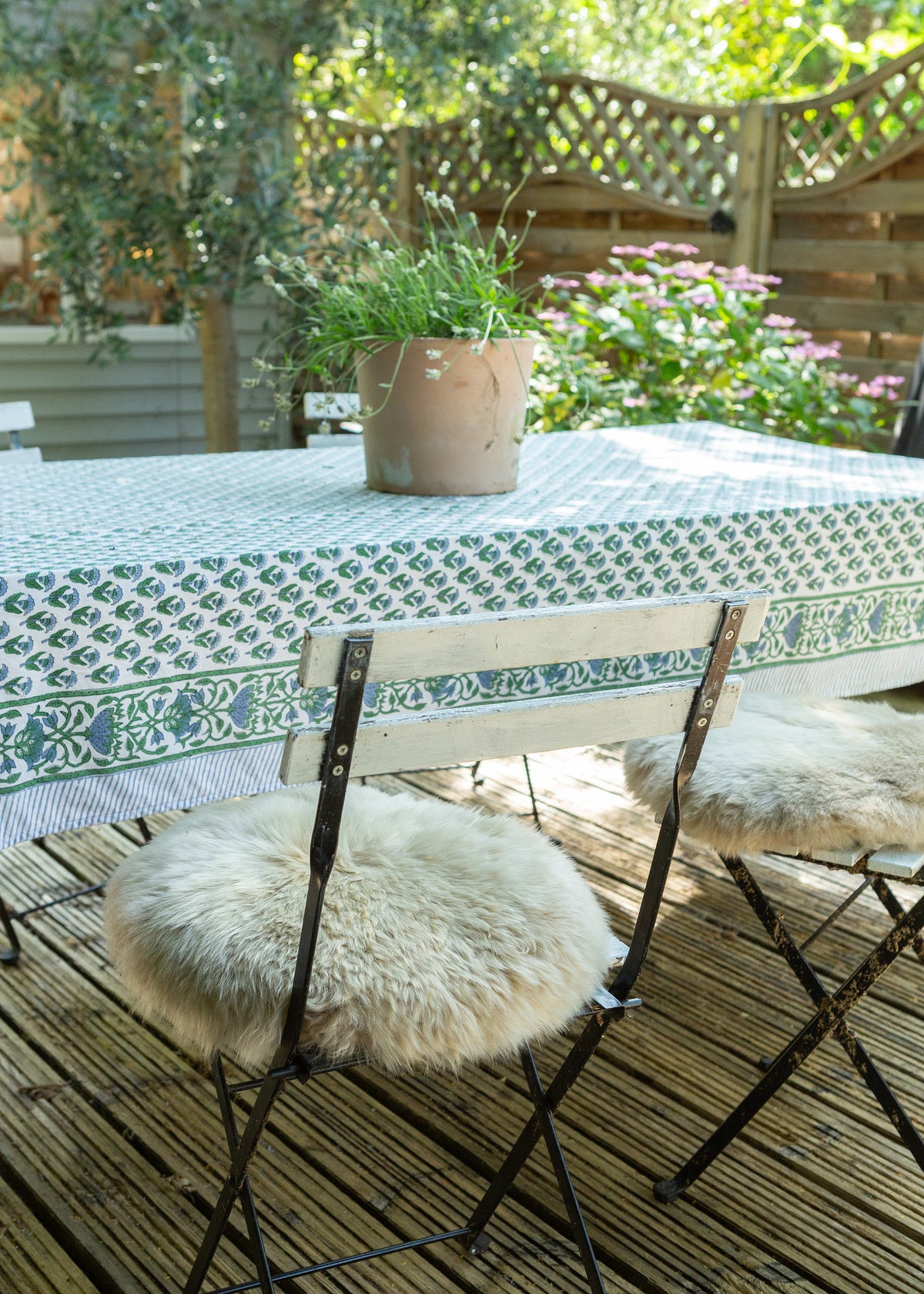 A styled outdoor dining scene featuring a rectangular table covered with a green and white patterned tablecloth, accompanied by two folding chairs with fluffy cream sheepskin seat cushions. A small potted plant sits on the table, set on a wooden deck with a garden background.